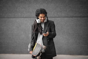 Nahaufnahme eines stilvollen, gutaussehenden glücklichen Dreadlocks-Skater-Mannes in einem Anzug, der mit Skate und Handy in den Händen steht und auf das Telefon schaut.