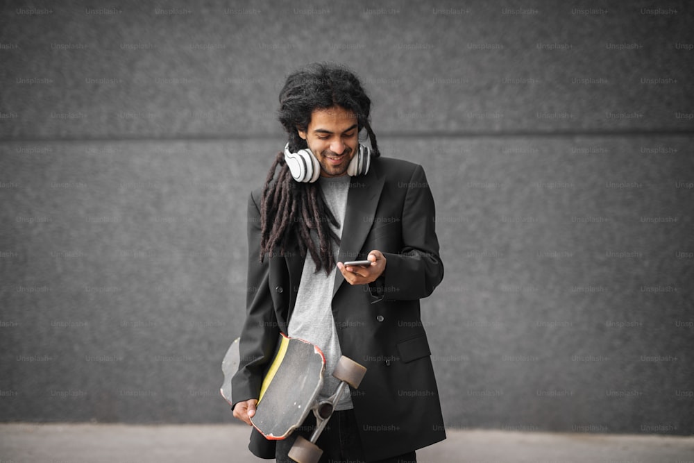 Close up of stylish handsome happy dreadlocks skater man in a suit standing with skate and mobile in hands and looking at the phone.
