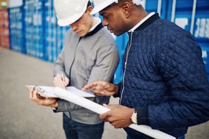 Two engineers wearing hardhats standing in a shipping container yard discussing an inventory list on a clipboard