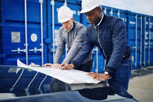 Two engineers leaning on the hood of a truck discussing blueprints while standing by freight containers on a commercial shipping dock