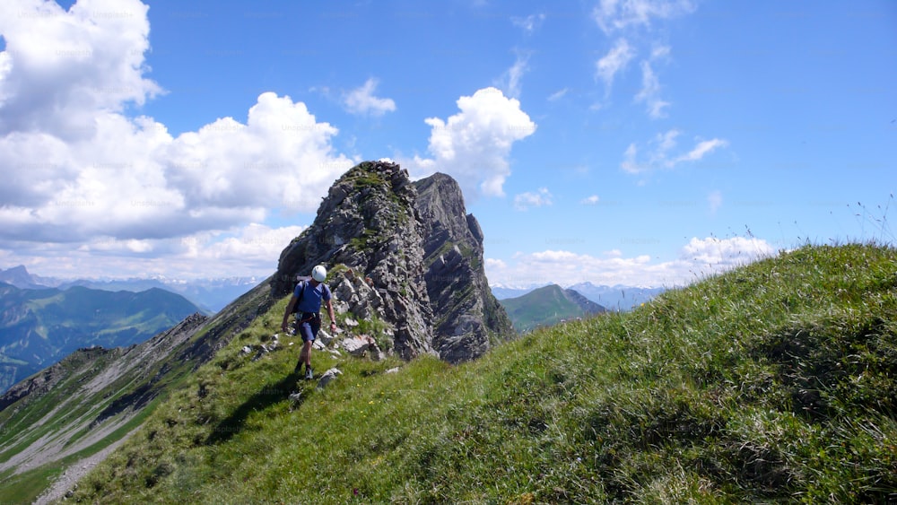A male climber on a grassy ridge on his way down from a climbing route with a great mountain landscape behind him
