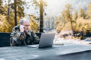 Serious pensive senior entrepreneur in work overalls is having a phone conversation while sitting at the wooden table with the laptop outdoors in the mountains in his summer place