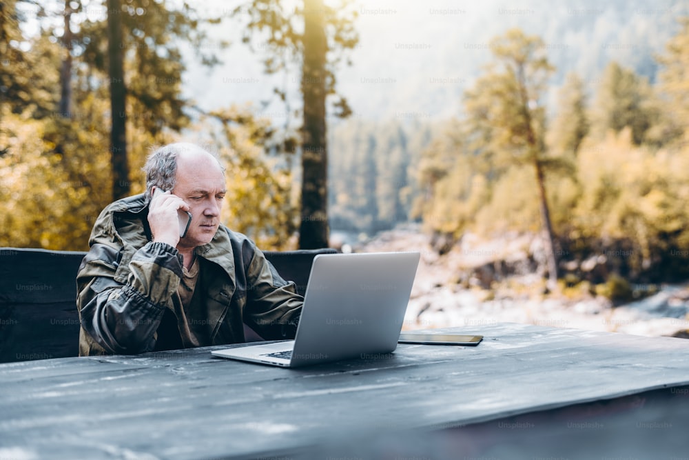 Serious pensive senior entrepreneur in work overalls is having a phone conversation while sitting at the wooden table with the laptop outdoors in the mountains in his summer place