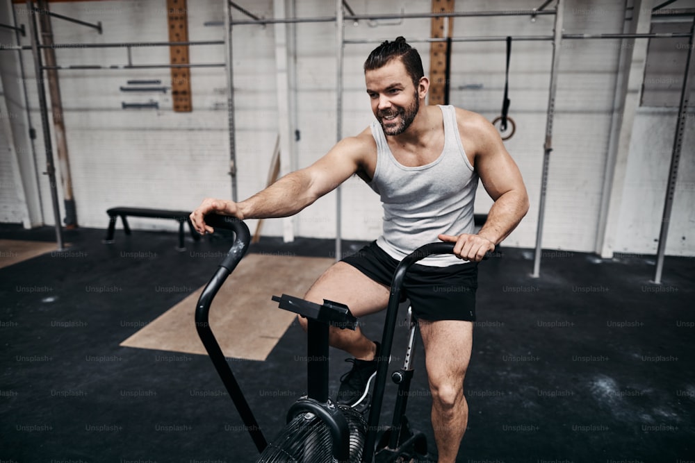 Hombre joven en forma en ropa deportiva sonriendo mientras suda montando una bicicleta estática durante una sesión de entrenamiento en el gimnasio