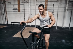 Jovem em forma em roupas esportivas sorrindo enquanto trabalha com suor andando de bicicleta ergométrica durante uma sessão de treino na academia