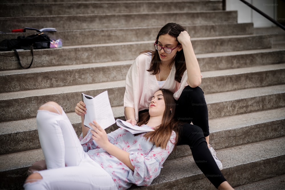 Pretty high school girl lying on stairs leaning against schoolmate girl leg and reading together school notes.