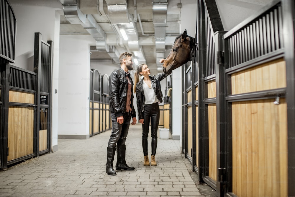 Young couple riders in leather jackets standing with horse in the beautiful stable