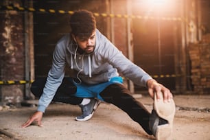 Retrato de cerca de un joven atlético atractivo afroamericano activo que hace un entrenamiento de estiramiento de piernas completas dentro del lugar abandonado.