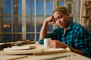 Tired female woodworker holding mug with aromatic hot beverage while sitting near workbench in workshop.