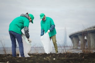Two guys in greenpeace uniform picking up litter into big sacks while workin outdoors
