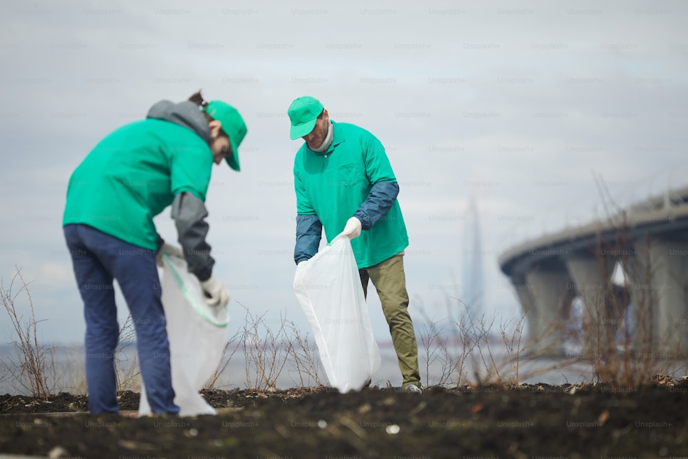 Two guys in greenpeace uniform picking up litter into big sacks while workin outdoors