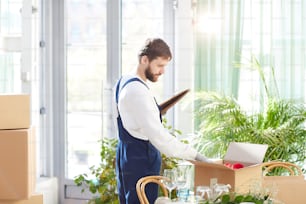 Serious concentrated handsome male moving company worker in uniform finding things while examining content of delivered boxes in new restaurant
