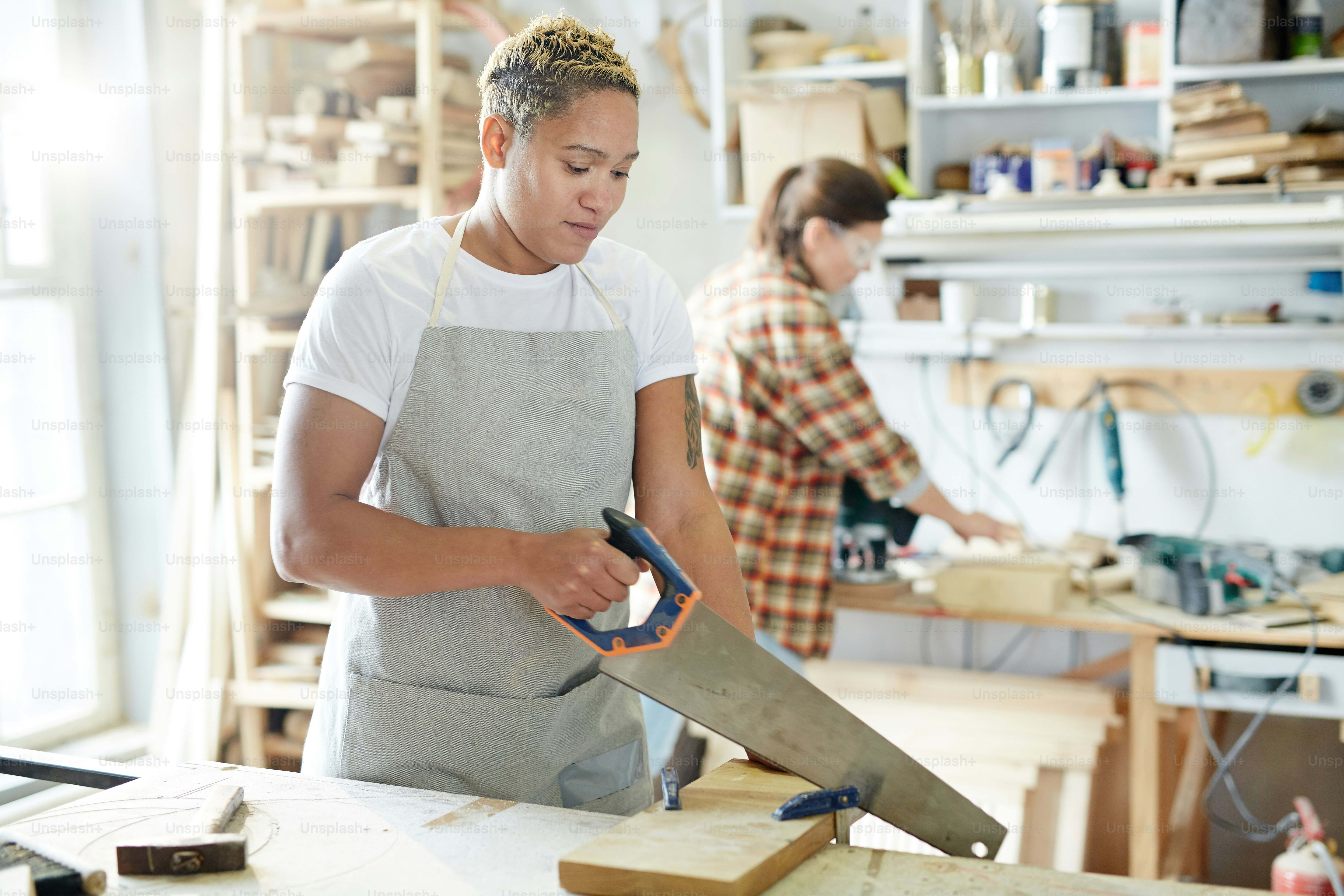 若い女性大工は、背景に彼女の同僚とワークショップで木の板を鋸で挽く