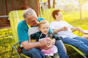 Elderly man sitting on a lazy chair outside holding his gorgeous smiling grandson and offering him water.