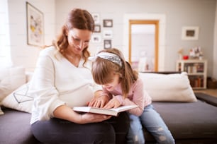 Pretty playful toddler girl sitting next to her careful pregnant mum and trying to read the book.