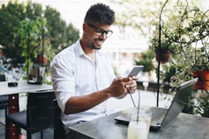 Joven y atractivo hombre de negocios afroamericano sentado en el bar de la cafetería haciendo un poco de trabajo en la computadora portátil y tomando en el teléfono móvil.