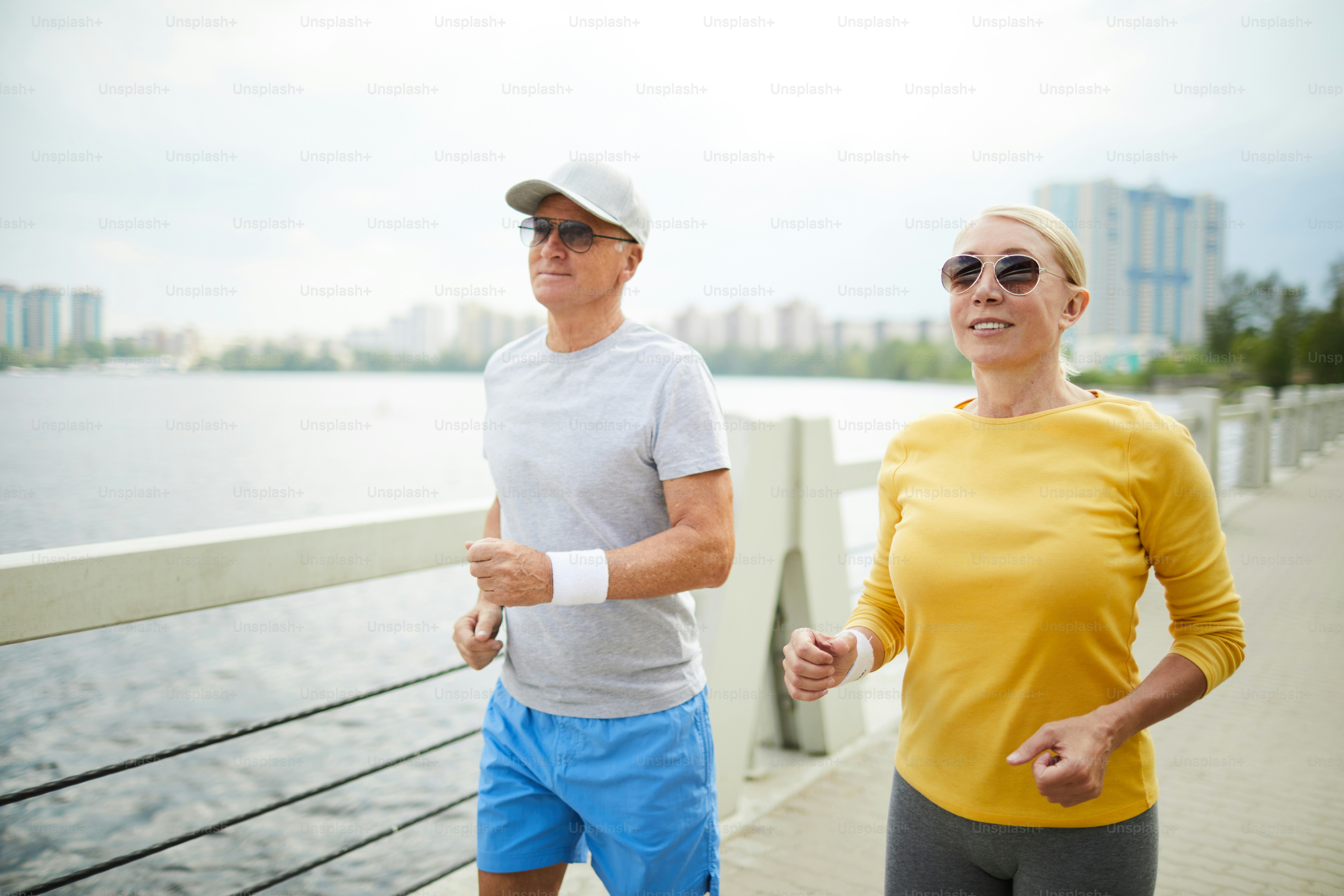 Two active seniors in sportswear and sunglasses jogging by waterside on summer morning