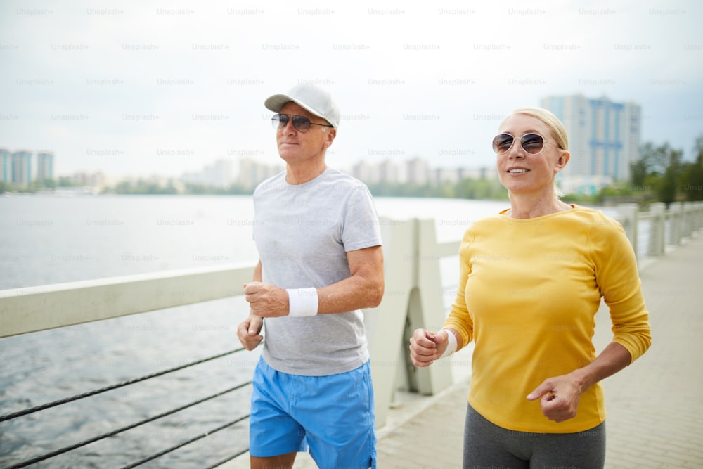 Dos personas mayores activas en ropa deportiva y gafas de sol trotando junto al agua en una mañana de verano
