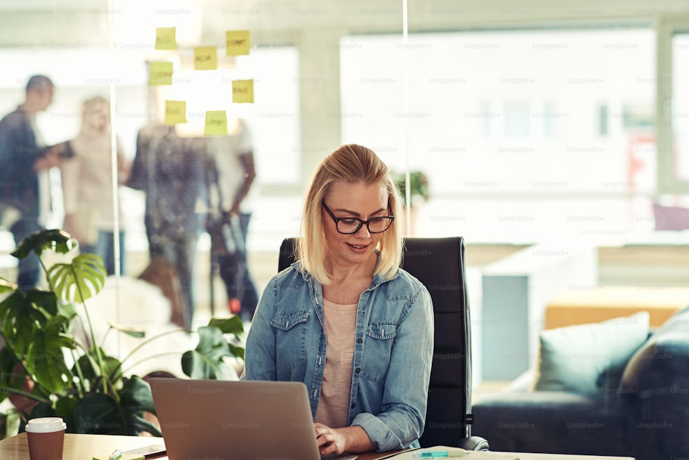 Casually dressed young businesswoman wearing glasses reading paperwork and working on a laptop while sitting at a desk in a modern office