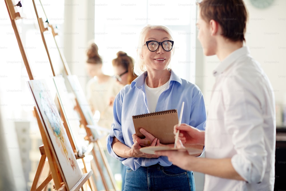 Confident teacher of arts talking to one of her students about professional painting traits