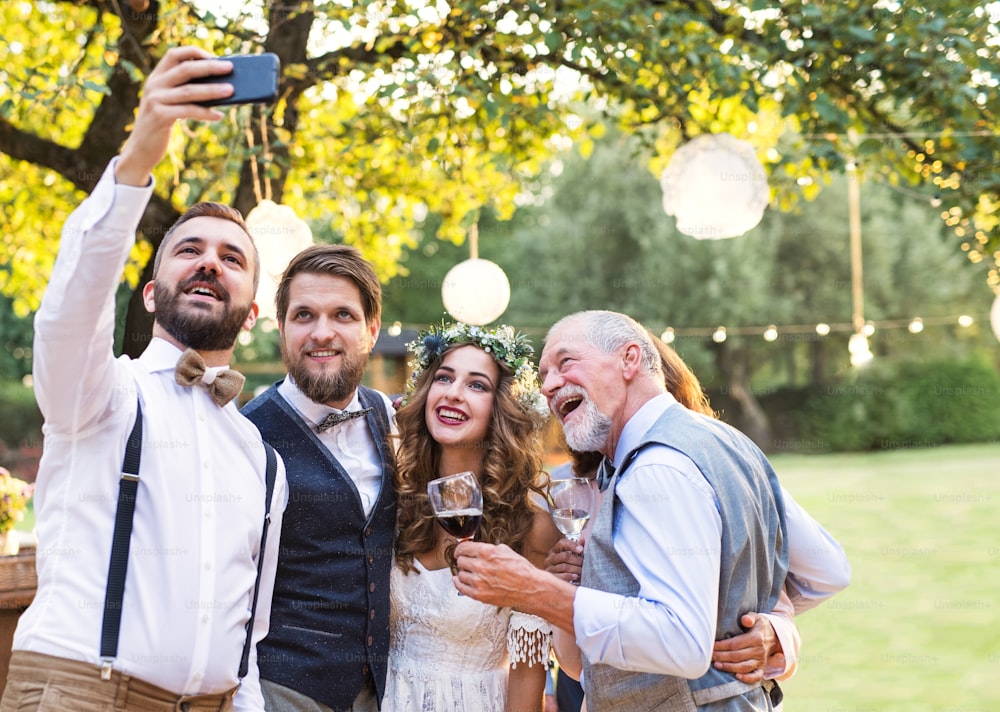 Happy bride, groom and guests with smartphone taking selfie outside at wedding reception.