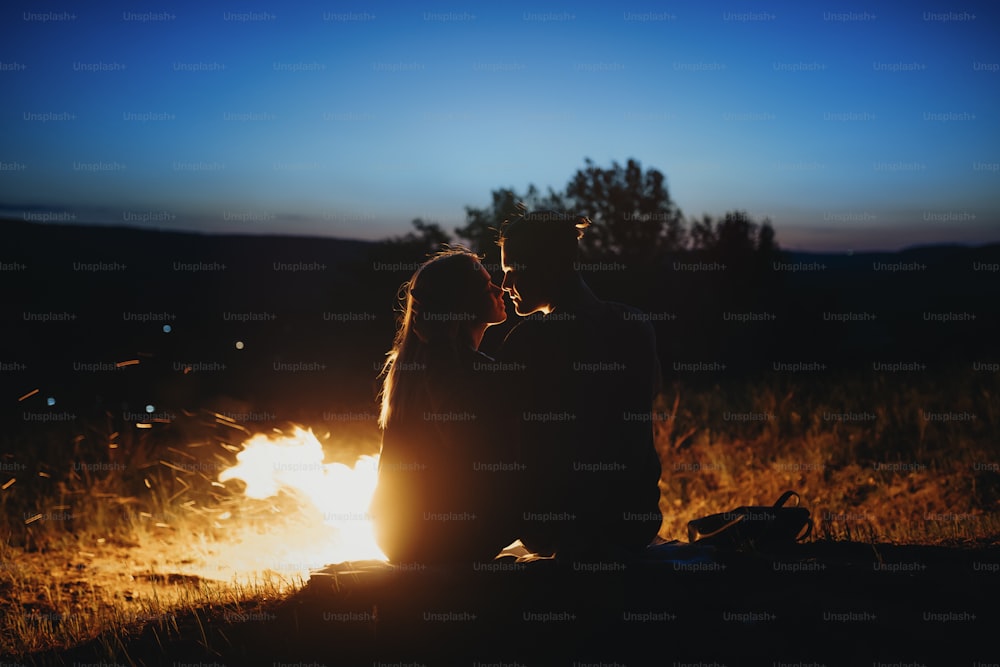 Retrato de vista lateral de una hermosa pareja joven sentada en el suelo cerca del fuego por la noche y mirándose el uno al otro viajando en su tiempo de vacaciones.
