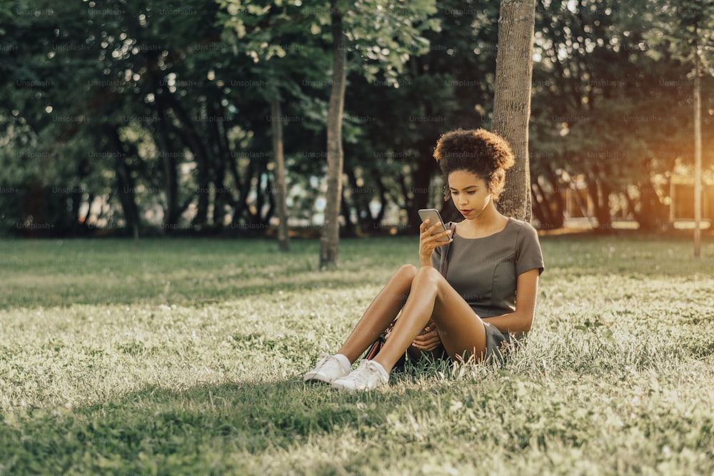 Giovane affascinante donna brasiliana con capelli afro ricci è seduta sul prato erboso nel parco pubblico, appoggiata all'albero e pubblicando i suoi selfie sui social network utilizzando lo smartphone