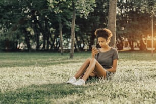 Young charming Brazilian female with curly Afro hair is sitting on the grass lawn in the public park, leaning against the tree and posting her selfies into social networks using the smartphone