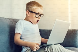 Momento sorprendente. Niño extremadamente sorprendido con gafas grandes mirando la pantalla de su nueva computadora portátil