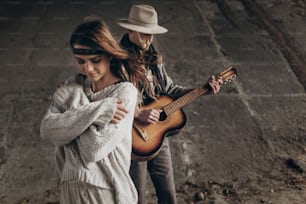 stylish hipster couple posing. man in hat playing guitar for his boho woman in knitted sweater. atmospheric sensual moment. rustic fashionable look.