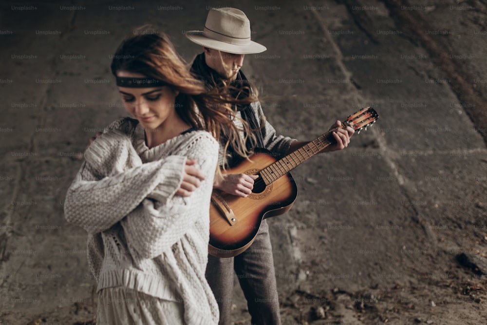 Stilvolles Hipster-Paar posiert. Mann mit Hut spielt Gitarre für seine Boho-Frau im Strickpullover. atmosphärischer sinnlicher Moment. rustikaler modischer Look.