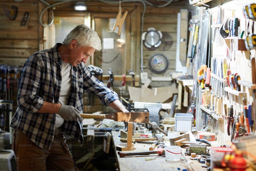 作業台のそばに立って木片を鋸で挽く作業服のシニア木工マスター