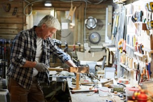 Senior woodworking master in workwear standing by workbench and sawing piece of wood