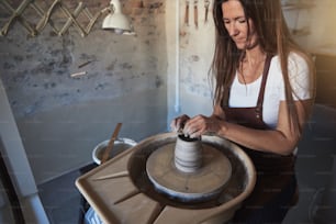 스튜디오의 도자기 바퀴에 앉아 젖은 점토 조각을 손으로 만드는 여성 장인