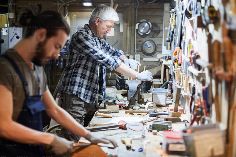 Reifer Meister der Holzbearbeitung mit Handwerkzeug zur Bearbeitung von Werkstücken mit seinem Auszubildenden in der Nähe