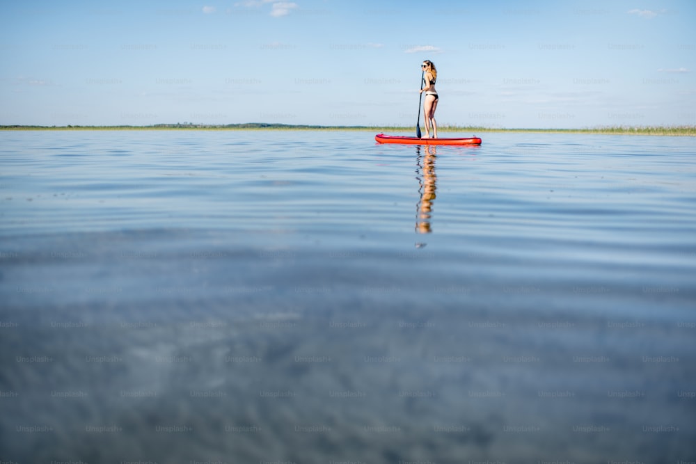 Giovane donna in costume da bagno nero paddleboarding sul lago con acqua calma e bella riflessione durante il tramonto. Ampia visuale con spazio di copia