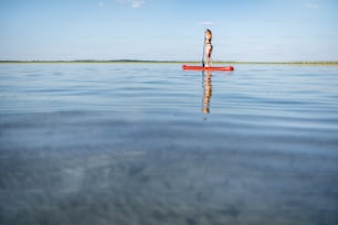 Young woman in black swimsuit paddleboarding on the lake with calm water and beautiful reflection during the sunset. Wide view with copy space