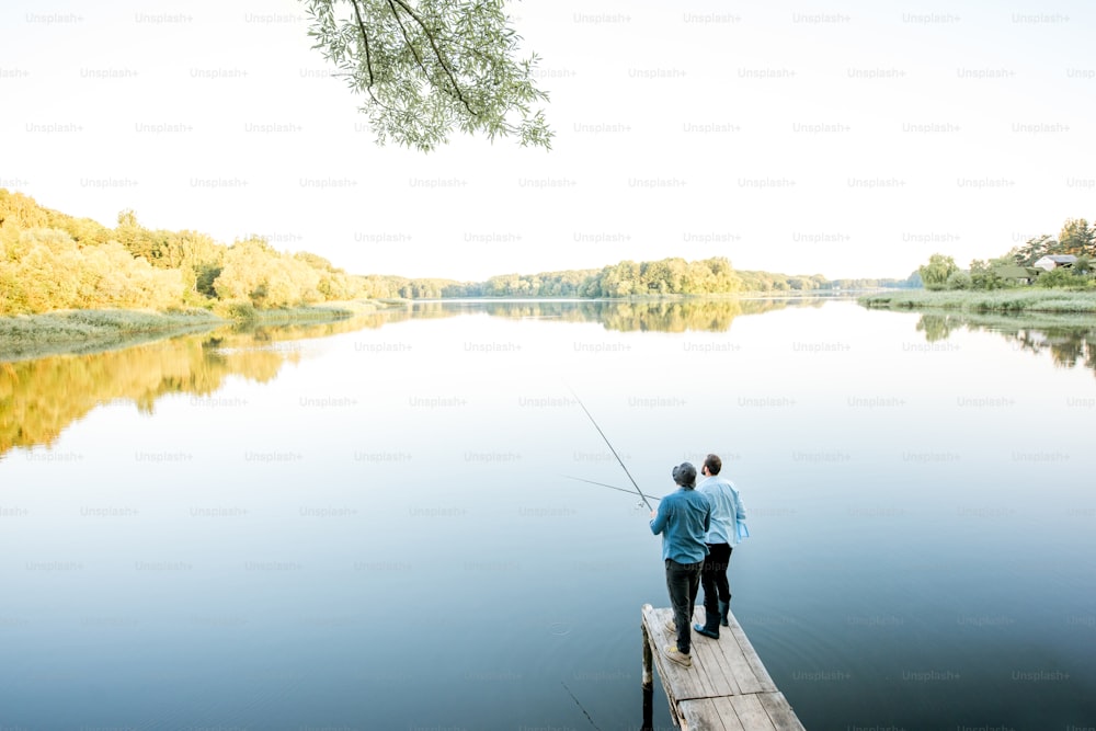 Landscape view on the beautiful lake with two male friends fishing together standing on the wooden pier during the morning light