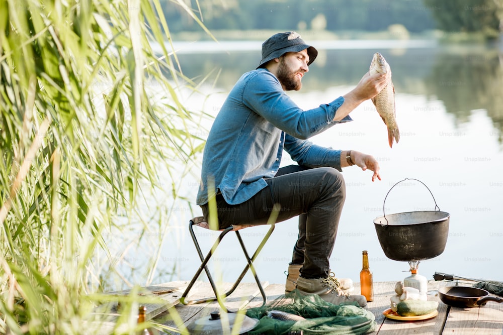 Pescador feliz sosteniendo pescado capturado listo para cocinar sentado durante el proceso de pesca cerca del lago