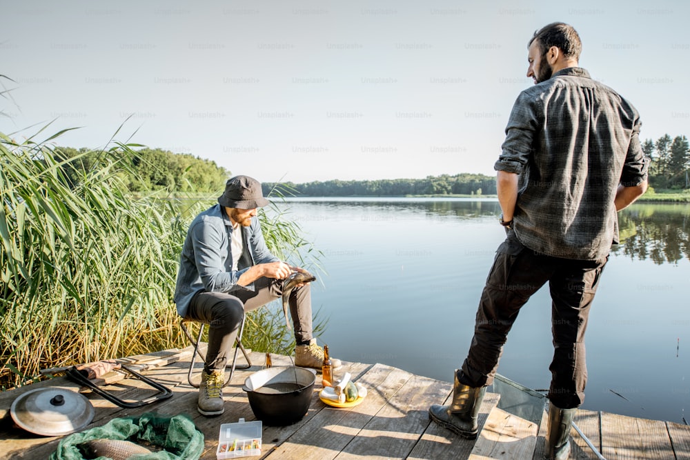 Two fishermen relaxing during the picnic on the wooden pier near the lake in the morning