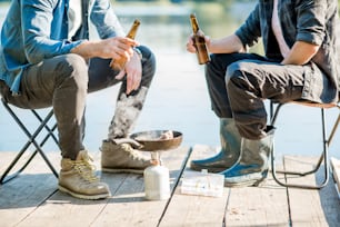 Dois pescadores fritando peixe sentados com cerveja durante o piquenique no cais de madeira perto do lago pela manhã