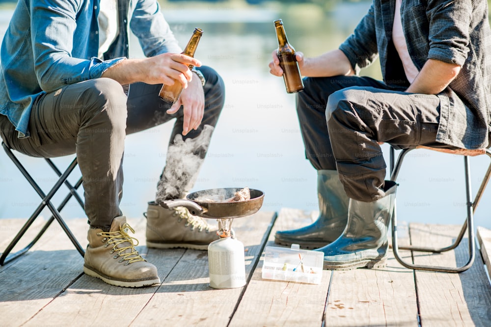 Two fishermen frying fish sitting with beer during the picnic on the wooden pier near the lake in the morning