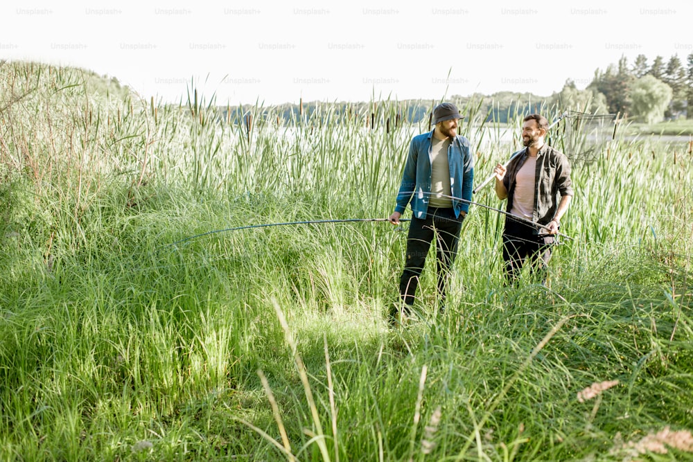 Two fishermen walking with fishing rod and net on the green lawn near the lake in the morning