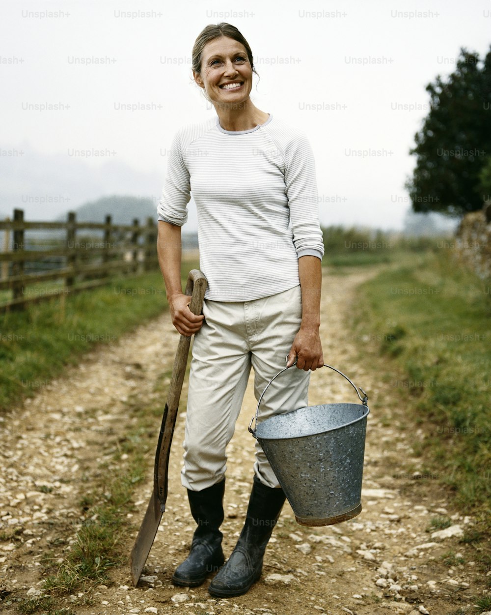 a woman standing on a dirt road holding a bucket and a shovel