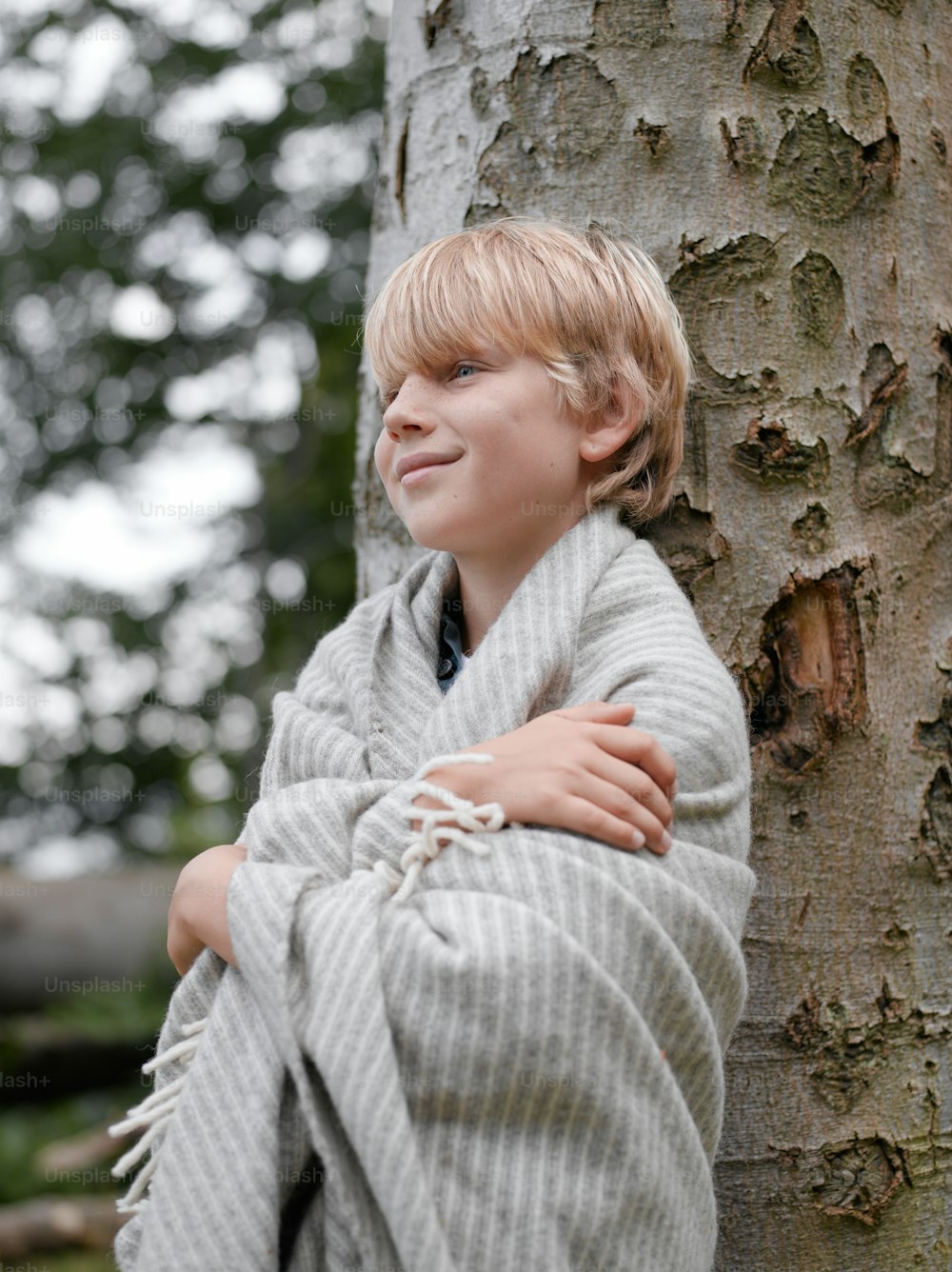 a young boy wrapped in a blanket leaning against a tree