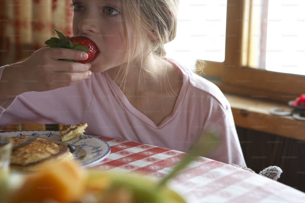 a little girl sitting at a table eating a strawberry