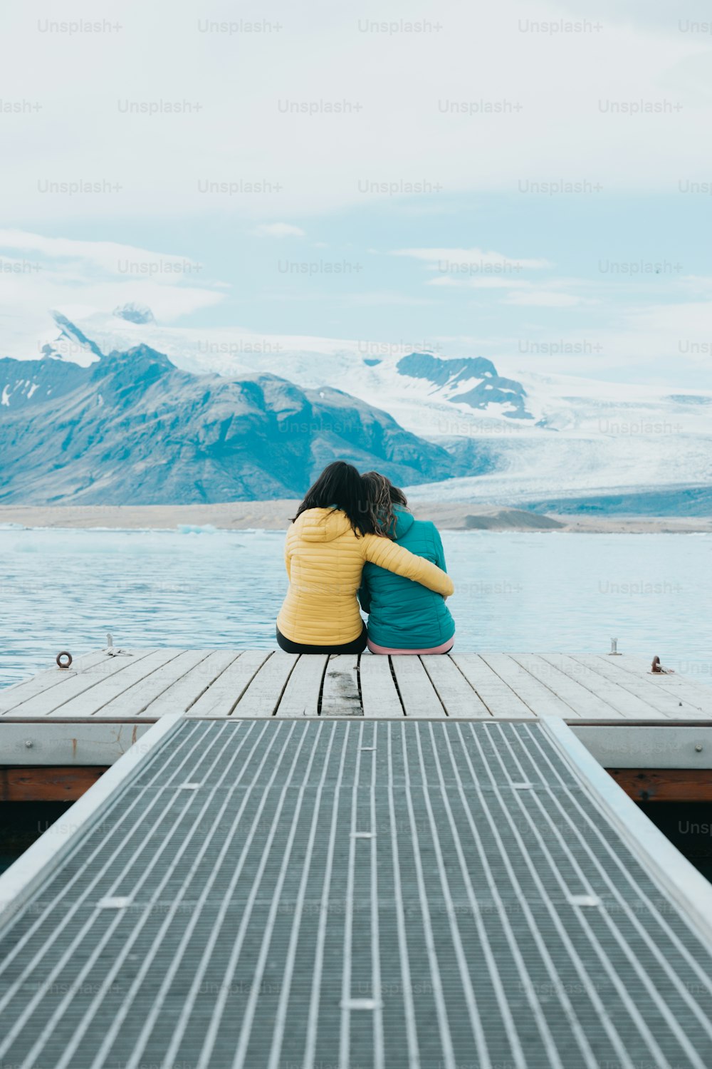 two people sitting on a dock with mountains in the background