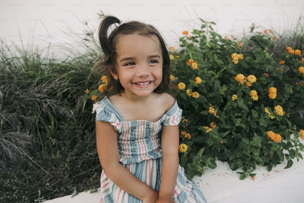 a little girl sitting on a ledge in front of flowers