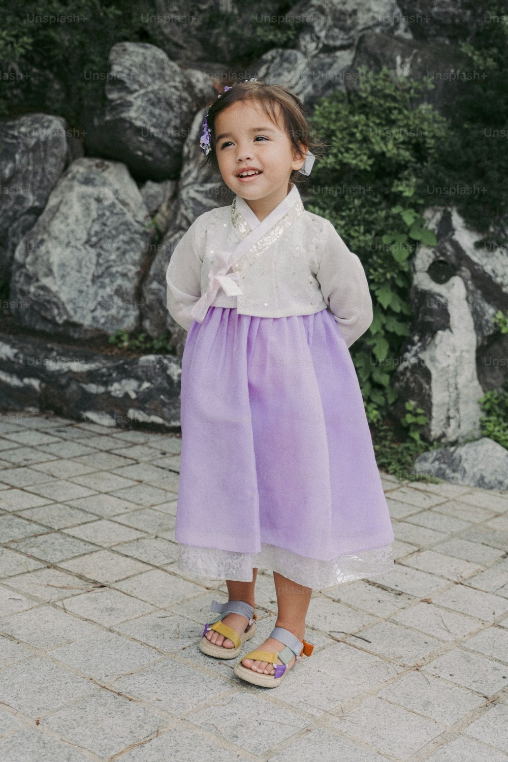 a little girl in a purple dress standing in front of some rocks
