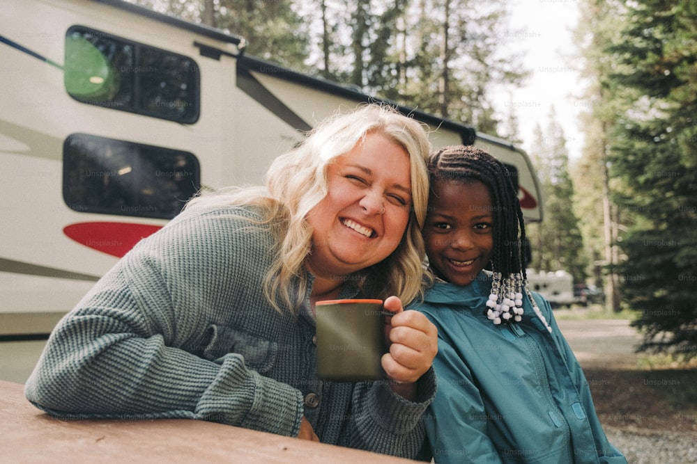 a woman holding a cup next to a girl in front of a camper